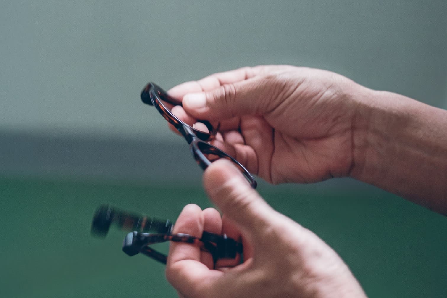 Two hands folding the temples of tortoiseshell glasses against a soft green background